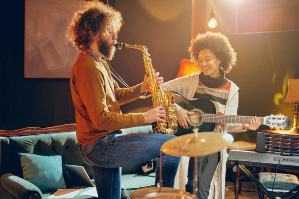 Mieszane rasy kobieta gra gitara akustyczna, podczas gdy człowiek grać na saksofonie. Domowym studio wnętrz. - Zdjęcie, obraz