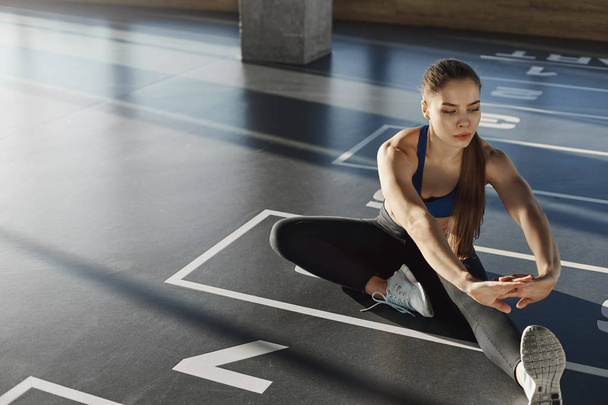 Focused woman athlete in sportswear sporty bra sitting gym floor stretching leg preparing muscles and biceps for working-out, pilates. Active young aerobics instructor practicing exercises - Photo, Image