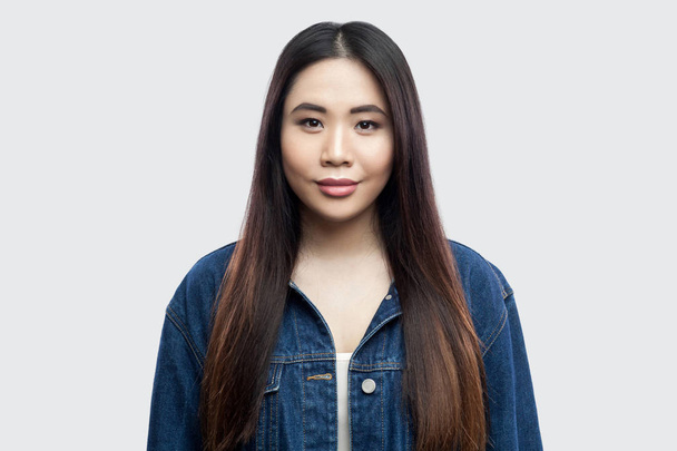 bela morena asiática jovem mulher no casual azul jaqueta de ganga com maquiagem sorrindo e olhando para a câmera no fundo cinza claro
. - Foto, Imagem