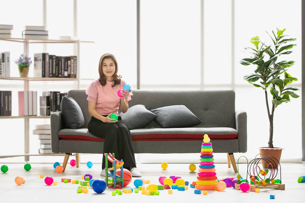 Asian young beautiful mother, happily smiling, sitting on sofa, holding multicolored balls in modern living room full of colorful toys scattered around on the floor - Photo, Image
