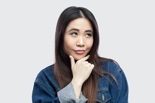 dreamy smiley beautiful brunette asian young woman in casual blue denim jacket touching her chin and pondering on light grey background. - Foto, immagini