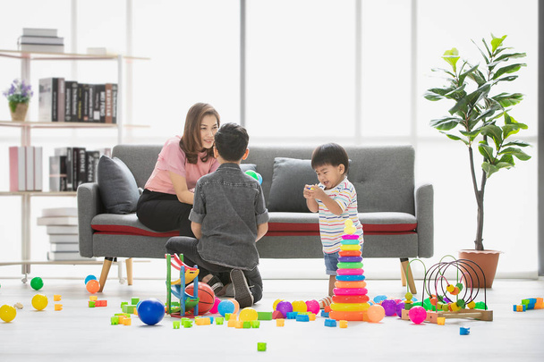 Asian family, happy time at home, young beautiful mother, taking care sons, little adorable toddler, standing, young boy wearing eyes glasses squat on floor, playing together in modern living room - Photo, Image