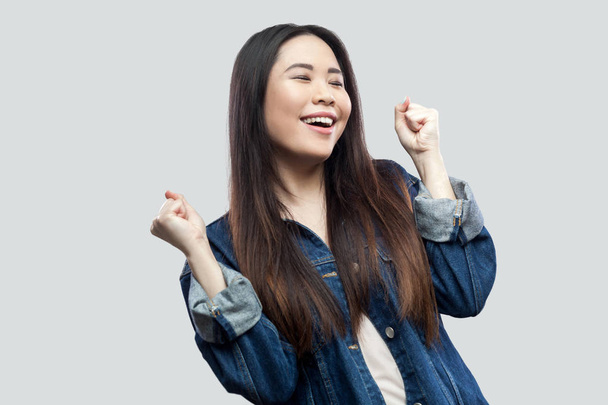 счастливый удивленный победитель красивая брюнетка азиатская молодая женщина в повседневной синей джинсовой куртке стоя и празднуя свою победу на светло-сером фоне
. - Фото, изображение