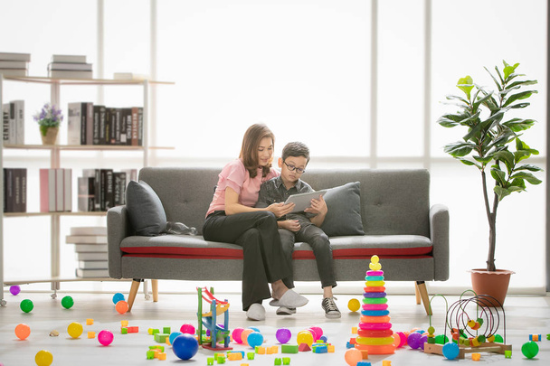 Asian family, happy, leisure time at home, young beautiful mother and young son wearing eyes glasses sitting on sofa, reading book, teaching homework in modern living room full of colorful toys - Photo, image