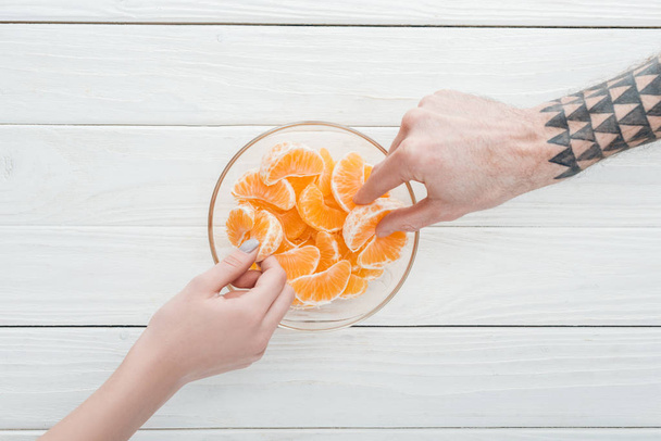 cropped view of couple taking tangerine slices from glass bowl on white wooden table - Фото, зображення