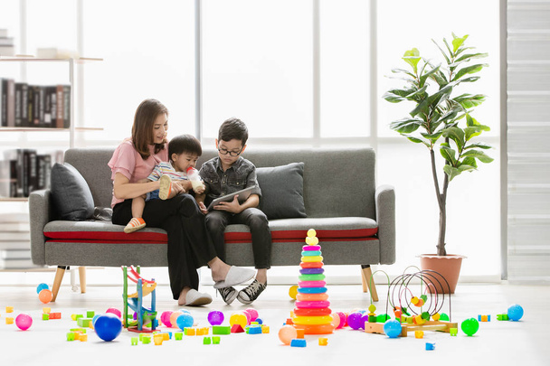 Asian family, happy, leisure time at home, young beautiful mother showing book to cute, little toddler drinking from milk bottle, young son wearing eyes glasses in her arms, in modern living room - Foto, immagini