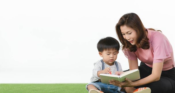 Ασιατική οικογένεια, ευτυχισμένος ελεύθερος χρόνος στο σπίτι, νεαρή όμορφη μητέρα, κάθεται σε τεχνητό γρασίδι, τρυφερά κρατώντας χαριτωμένο, αξιολάτρευτο μικρό παιδί στην αγκαλιά της, διαβάζοντας βιβλίο γι 'αυτόν, σε λευκό φόντο - Φωτογραφία, εικόνα