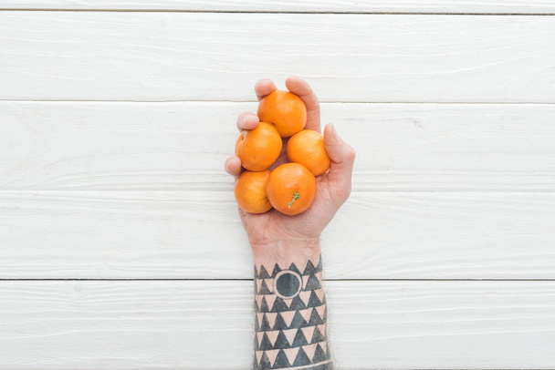 vista cortada do homem tatuado segurando montão de tangerinas na mão na superfície de madeira branca
 - Foto, Imagem