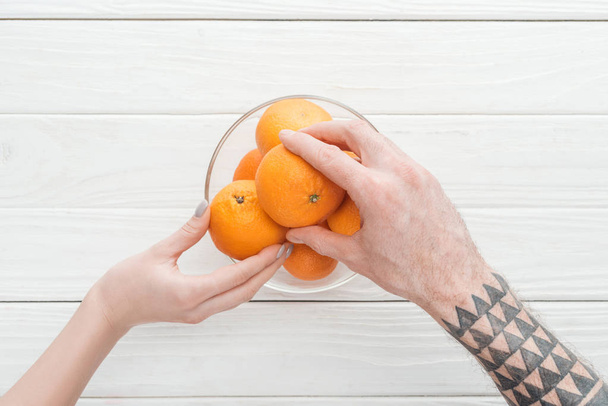 partial view of tattooed man and woman taking tangerines from glass bowl on white wooden surface - Photo, Image