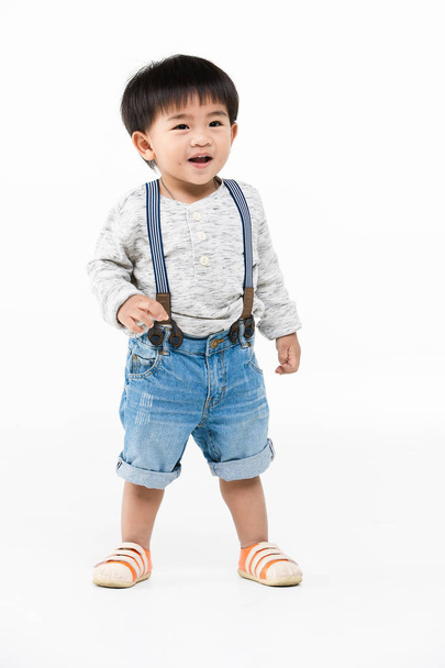 Studio portrait of cute, adorable, Asian toddler boy wearing denim overalls, long sleeve T-shirt, orange shoes, standing, smiling, hold his brace suspenders on isolated white background - Фото, изображение