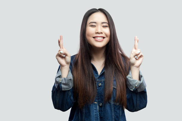 hopeful beautiful brunette asian young woman in casual blue denim jacket with makeup standing with crossed fingers and praying on light grey background. - Foto, immagini