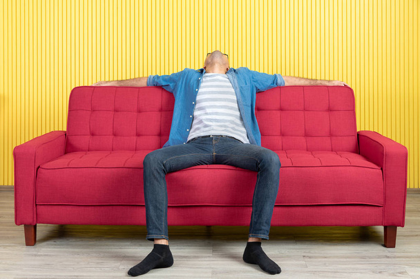 Young Asian handsome bearded man, wearing eyeglasses in denim shirt, sitting on center of red sofa, arms wide spread in confident, in modern living room, bright yellow stripe wall background - Foto, Bild