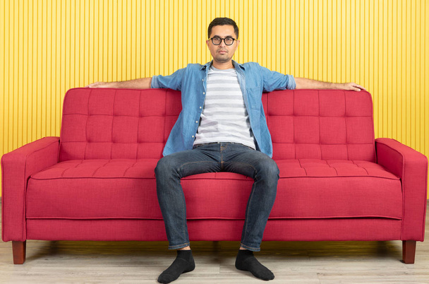 Giovane asiatico bell'uomo barbuto, indossando occhiali in camicia di jeans, seduto al centro del divano rosso, braccia largamente diffuse in fiducioso, in soggiorno moderno, luminoso sfondo della parete a strisce gialle
 - Foto, immagini