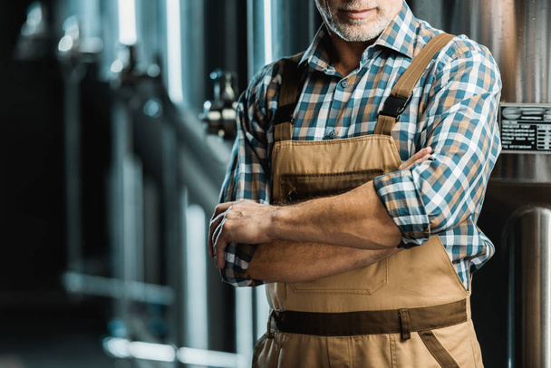 cropped view of brewer posing with crossed arms in working overalls in brewery - Photo, Image