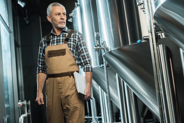 senior brewer in working overalls holding notepad while examining brewery - Photo, Image