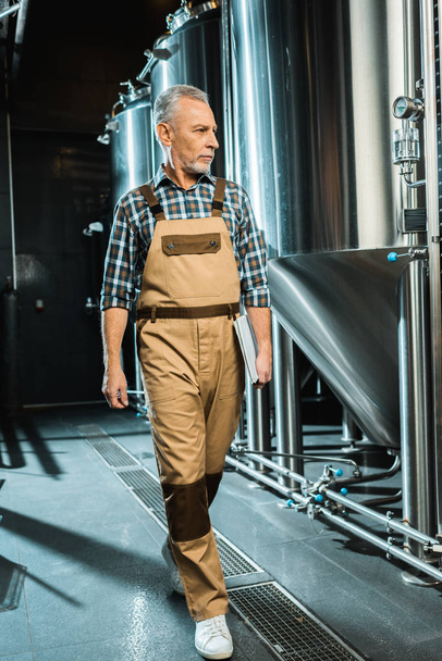 senior brewer in working overalls holding notepad while walking and examining brewery - Photo, Image