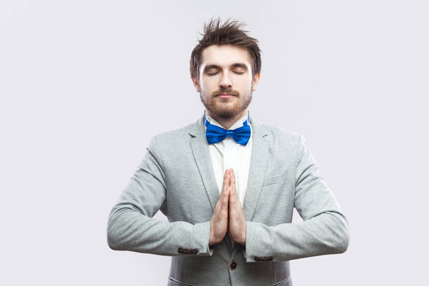calm relax handsome bearded man in casual grey suit and blue bow tie standing with closed eyes and doing yoga with palm hands while meditating on light grey background. - Zdjęcie, obraz