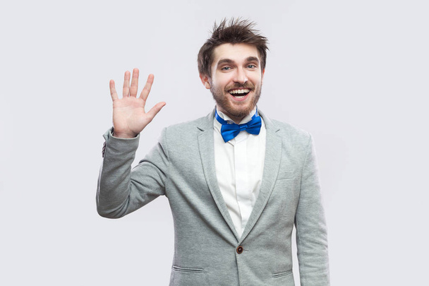 excited happy handsome bearded man in casual grey suit and blue bow tie standing and looking at camera, greeting and toothy smile. indoor studio shot, isolated on light grey background - Fotografie, Obrázek