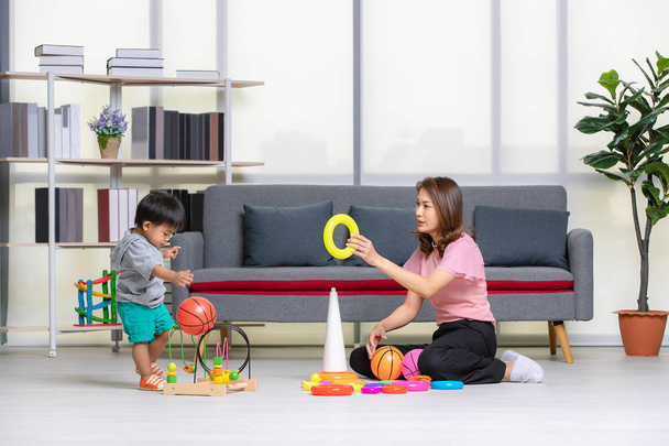 Happy Asian family, young, beautiful mother in pink blouse, sitting on floor, hold yellow ring, adorable little toddler boy in grey T-shirt, short, enjoy playing with ball and toys in cozy living room - Fotó, kép