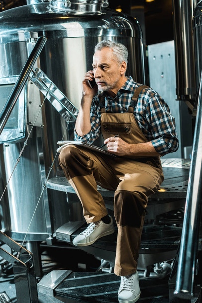 serious professional brewer in working overalls talking on smartphone while writing in notepad in brewery - Photo, Image