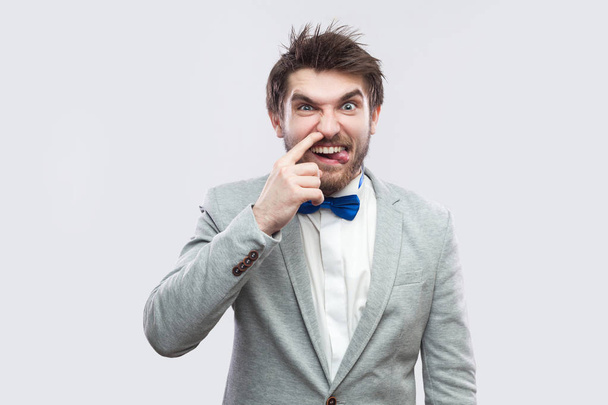 Portrait of funny handsome bearded man in casual grey suit and blue bow tie looking at camera with tongue out and drilling nose on light grey background. - Foto, imagen