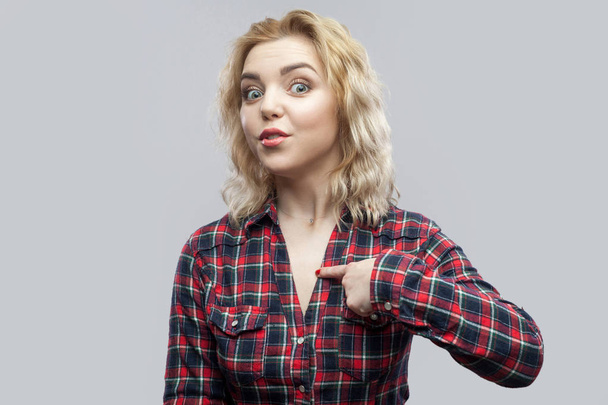 surprised beautiful blonde young woman in casual red checkered shirt standing and pointing herself with amazed face and asking on grey background - Φωτογραφία, εικόνα