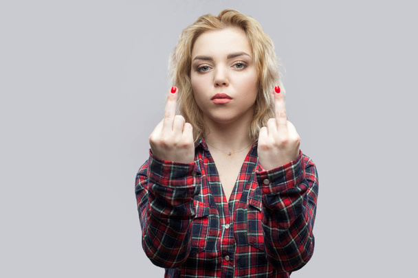 serious young beautiful blonde woman in casual red checkered shirt standing and looking with poker face and middle finger fuck signs on grey background. - Photo, image