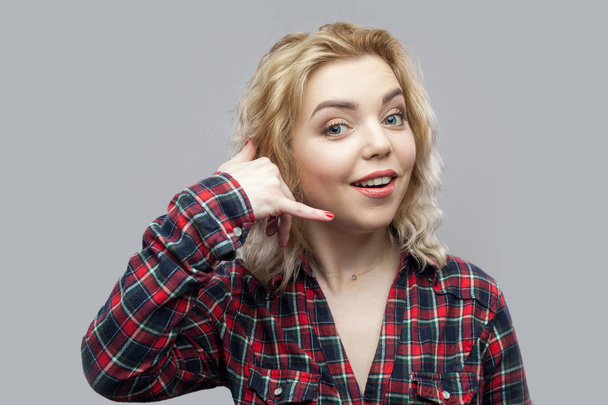 funny beautiful blonde young woman in casual red checkered shirt standing with call gesture and looking at camera while toothy smile on grey background. - Φωτογραφία, εικόνα