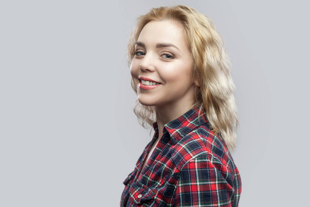 happy beautiful blonde young woman in casual red checkered shirt standing and looking at camera with toothy smile on grey background. - 写真・画像