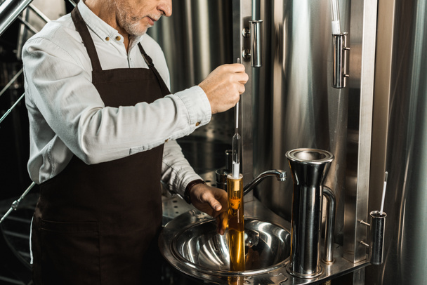 cropped view of senior brewer examining beer in flask in brewery - Photo, Image