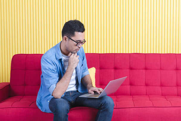 Fiatal ázsiai jóképű szakállas férfi, visel szemüveg farmering, ülő szélén a piros kanapé, böngészés weboldalak notebook modern nappali, fényes sárga csík fal háttér - Fotó, kép