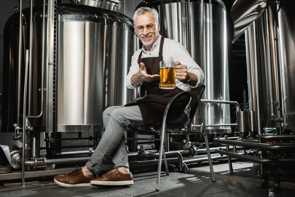 lachende senior brouwer in schort zittend op een stoel en glas bier in brouwerij tonen - Foto, afbeelding