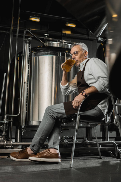Braumeister in Schürze sitzt auf Stuhl und testet Bier in Brauerei - Foto, Bild