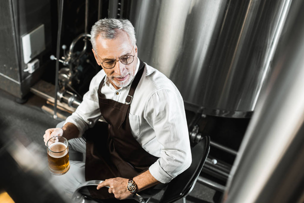 ogólny widok starszy piwowara w fartuch, siedząc na krześle i trzymając szklankę piwa w browarze - Zdjęcie, obraz