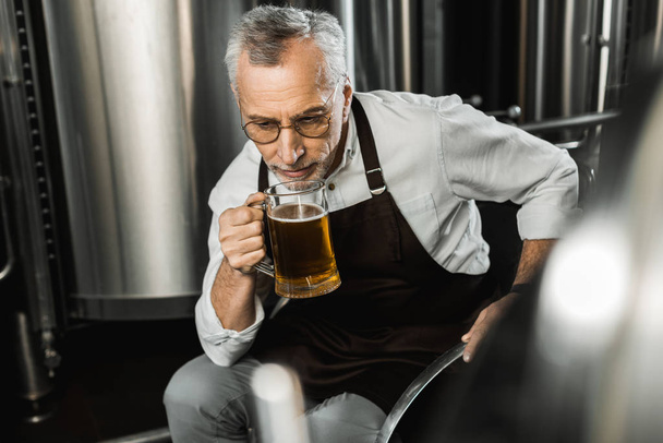 senior brewer in apron sitting on chair and testing beer in brewery - Φωτογραφία, εικόνα