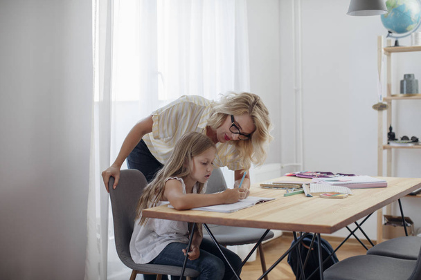 Beautiful blonde Caucasian woman helping to her cute daughter to study at home. - Foto, afbeelding