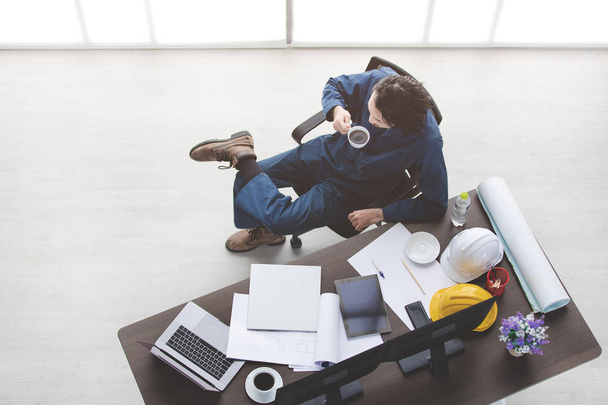 Top view of Asian beard male foreman, in coverall, wear safety shoes, relax, sit on chair, coffee cup on table, with stationery, safety helmets, laptop, tablet, smartphone, office background - Foto, Imagen