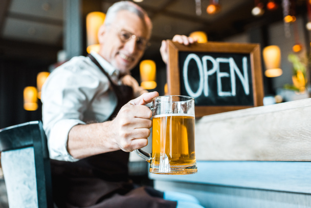 focalizzazione selettiva di proprietario anziano di pub che tiene il segno aperto e il vetro di birra
 - Foto, immagini