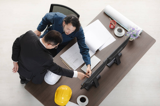 Draufsicht asiatischer Geschäftsmann zeigt auf Desktop-Bildschirm auf asiatischen männlichen Vorarbeiter in Overall, sitzt auf Stuhl, diskutiert über neues Projekt, mit Bauplan und Helmen und Bürohintergrund - Foto, Bild