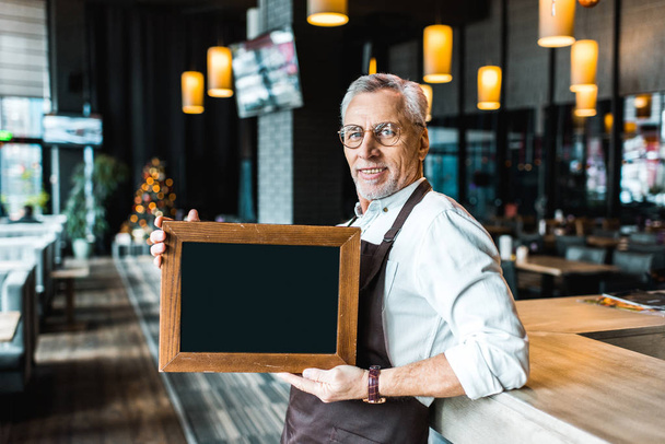 smiling owner of pub holding empty board and standing near bar counter  - Photo, Image
