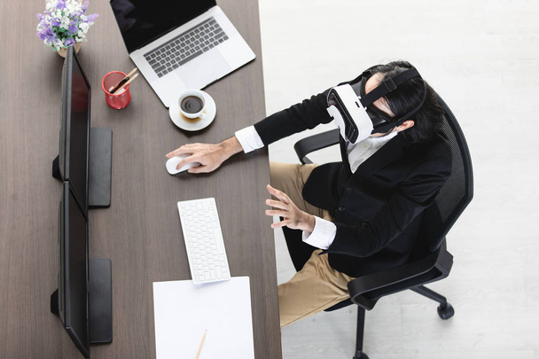 Вид сверху на молодого азиатского бизнесмена в очках виртуальной реальности, протягивающего обе руки перед собой для тестирования видения, занятого просмотром нового гаджета, с ноутбуком, современным офисным фоном
 - Фото, изображение