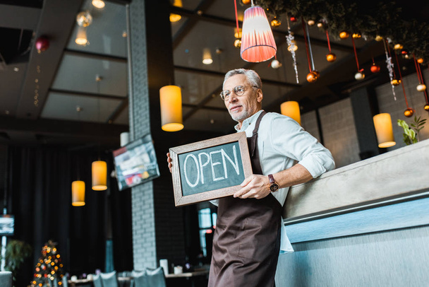 senior worker of pub holding open sign and standing near bar counter  - Photo, Image