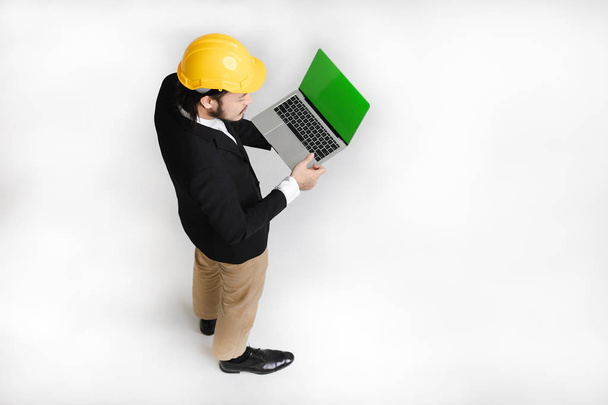 Top view of happy, young Asian business man in black suit and white shirt, wearing yellow helmet, looking down and touching at tablet screen in hand, on white isolated background - Photo, Image