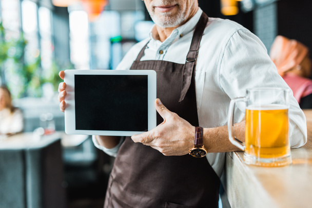 vista recortada del propietario masculino sosteniendo tableta digital con pantalla en blanco en el pub con vaso de cerveza
 - Foto, Imagen