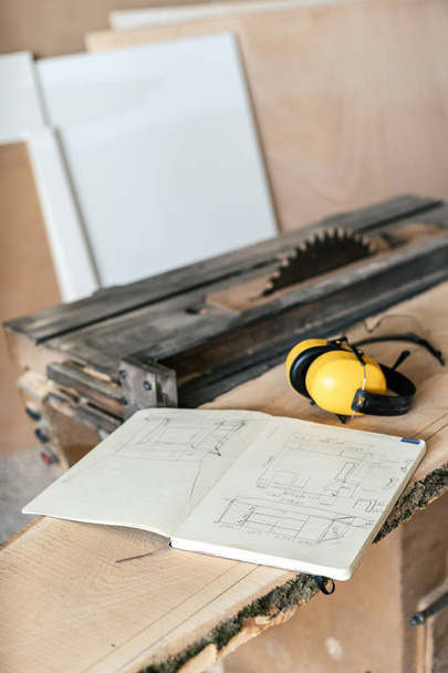 Vertical photo of notebook with technical drawing lying on top of wood plank board in garage or workroom near yellow headset and table saw - Foto, Imagen