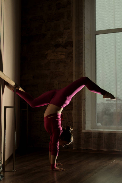 Όμορφο κορίτσι χορευτής σε ένα ροζ κοστούμι κάνει προθέρμανση barre το μπαλέτο. προθέρμανση σπορ στην αίθουσα χορού. Διάφορες πόζες και ασκήσεις κοντά το barre μπαλέτο. - Φωτογραφία, εικόνα
