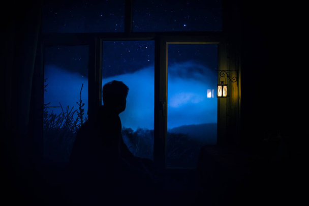 Silhouette of a man looking a dreamlike galaxy through a window. Fantasy picture with old vintage lantern at the window inside dark room. - Photo, Image