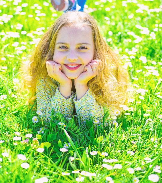 Heyday concept. Girl on smiling face spend leisure outdoors. Girl lying on grass at grassplot, green background. Child enjoy spring sunny weather while lying at meadow - 写真・画像