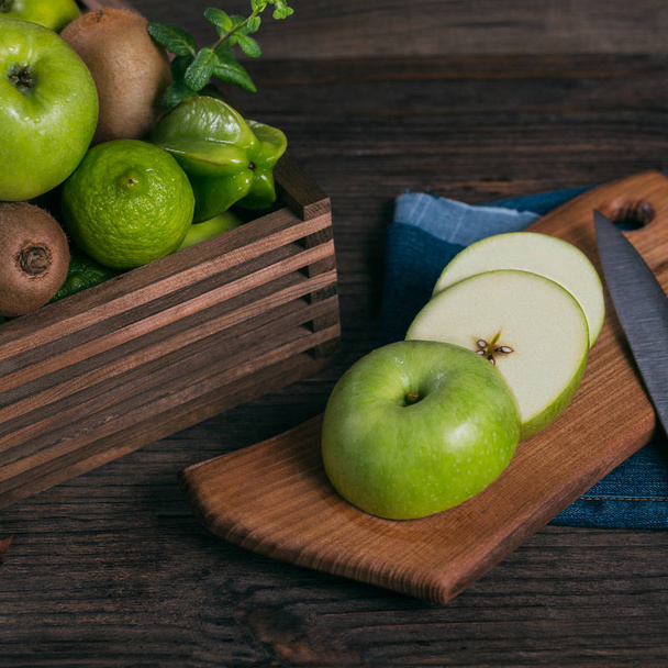 Concept of healthy food. Fresh green fruits in a box, green apple on cutting board. Dark wooden background. Set of green fruits for healthy diet and detox: apple, lime, kiwi, mango, carambola and mint - Zdjęcie, obraz