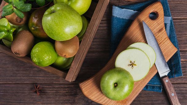 Concept of healthy food. Fresh green fruits in a box, green apple on cutting board. Dark wooden background. Set of green fruits for healthy diet and detox: apple, lime, kiwi, mango, carambola and mint - Фото, зображення